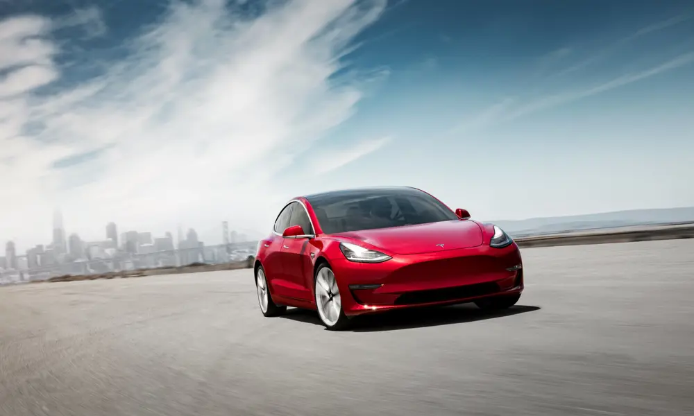 A red Model 3 Tesla driving on a road with a city in the background.