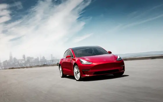A red Model 3 Tesla driving on a road with a city in the background.