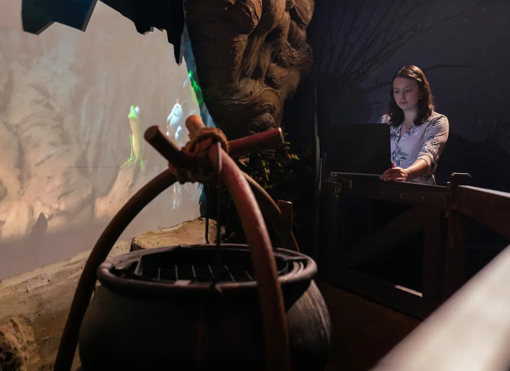 A replica cauldron is in a dark room. Behind it is a screen showing an animation of a frog and a dog, and next to that is a woman in a white shirt working at a computer