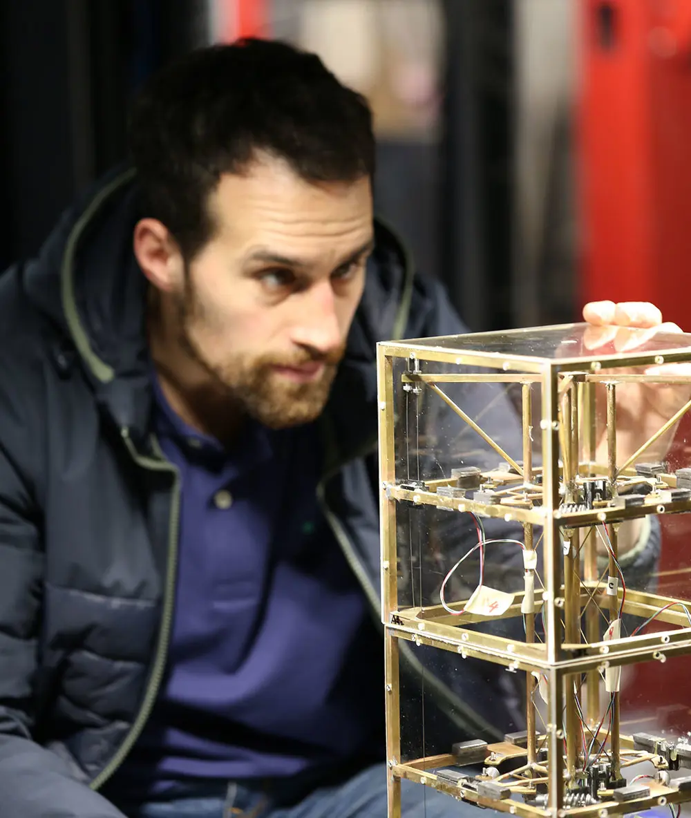 A person examines a small cube-shaped mechanical structure with visible wiring and metal supports.