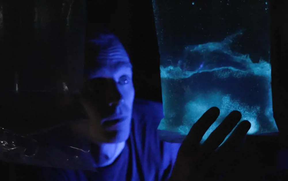 In the dark, a man holds up a clear plastic bag full of glowing algae, which illuminates his face