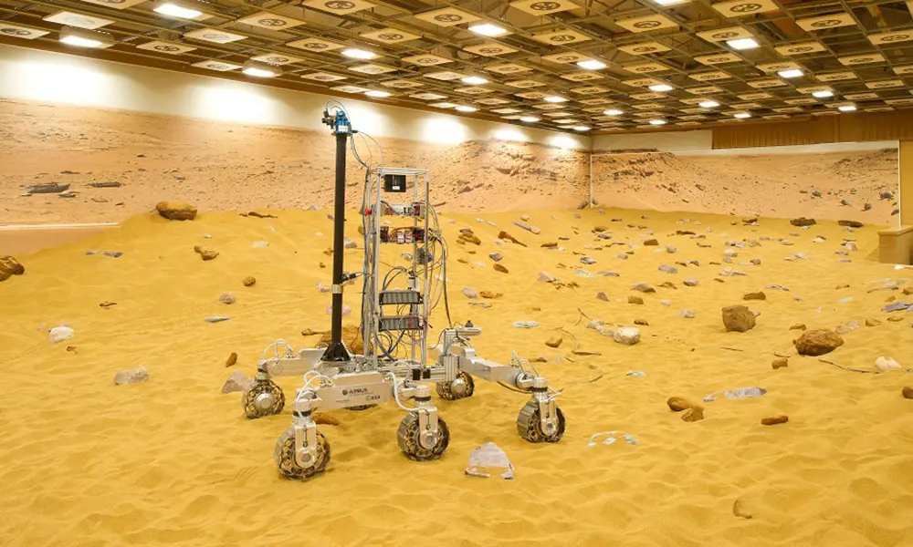 The ExoMars rover at the Stevenage Airbus aerospace premises. It is placed in a sandpit that is covered with rocks in a room full of lights. 