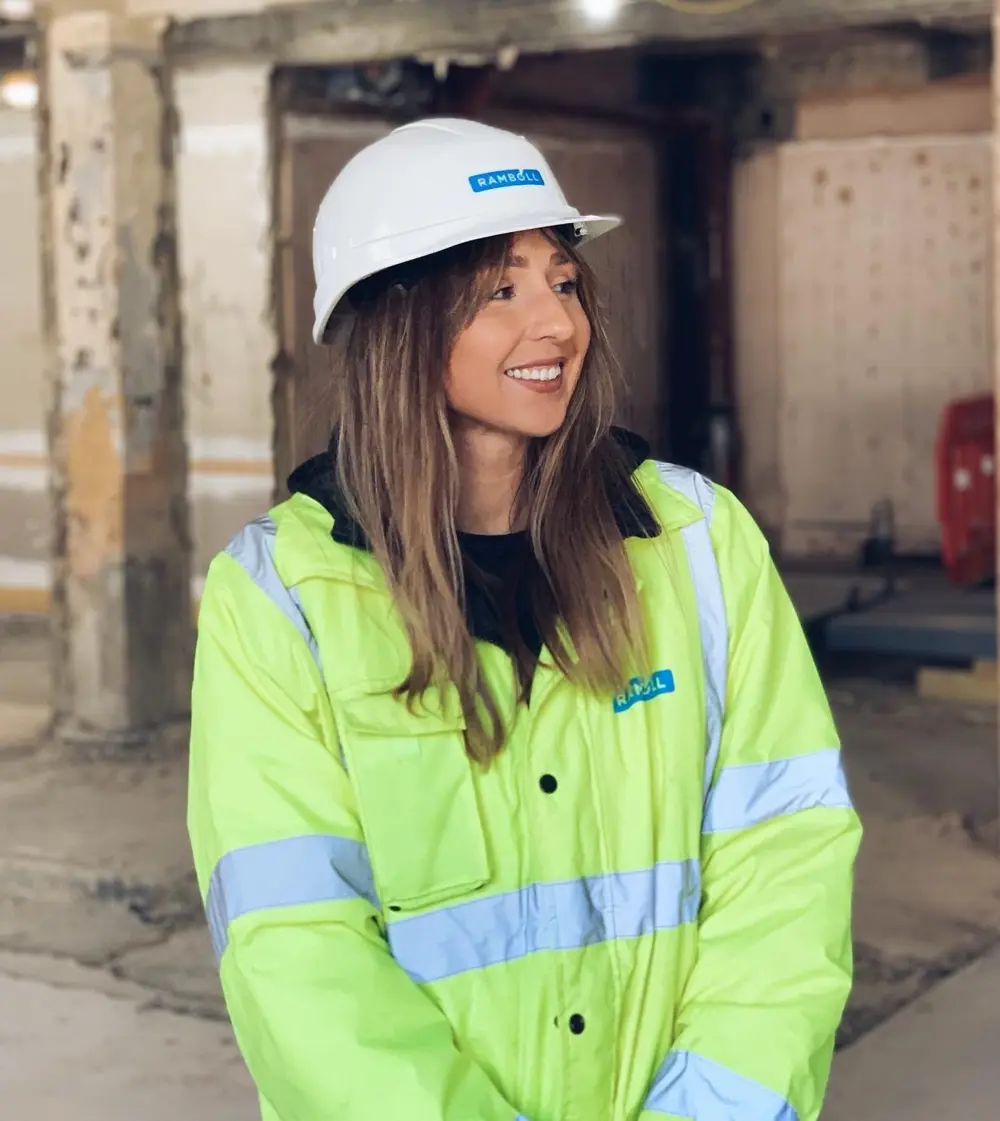 Person in a high-visibility jacket and safety helmet standing on a construction site, with industrial structures in the background.