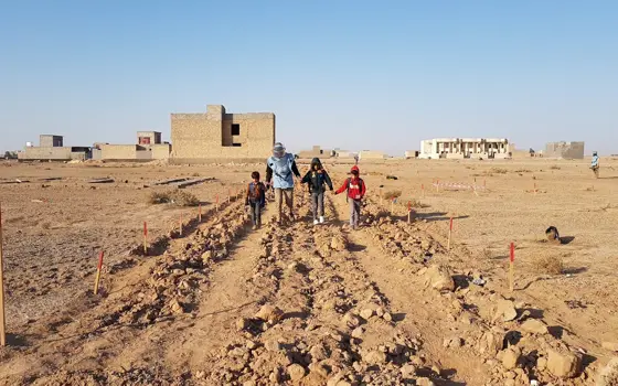 Three children and an adult in a blue vest holding hands as they cross a path demarcated across an arid field.
