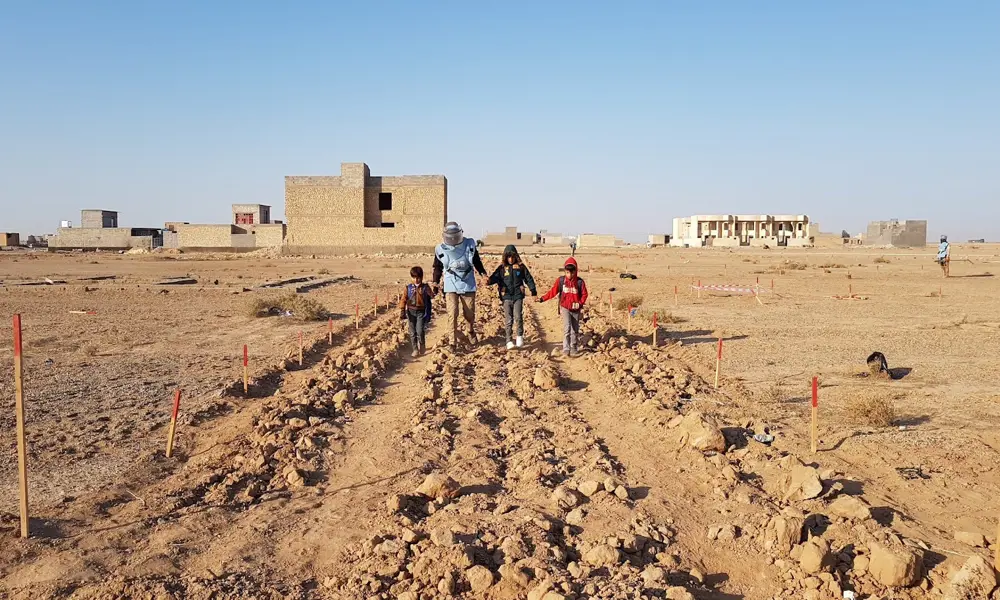 Three children and an adult in a blue vest holding hands as they cross a path demarcated across an arid field.