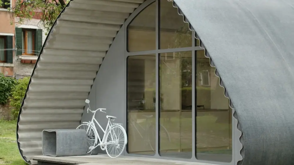 Close-up of the Essential Home prototype, showing a curved shell made from rollable concrete material, large glazed entrance panels and a bicycle parked outside.