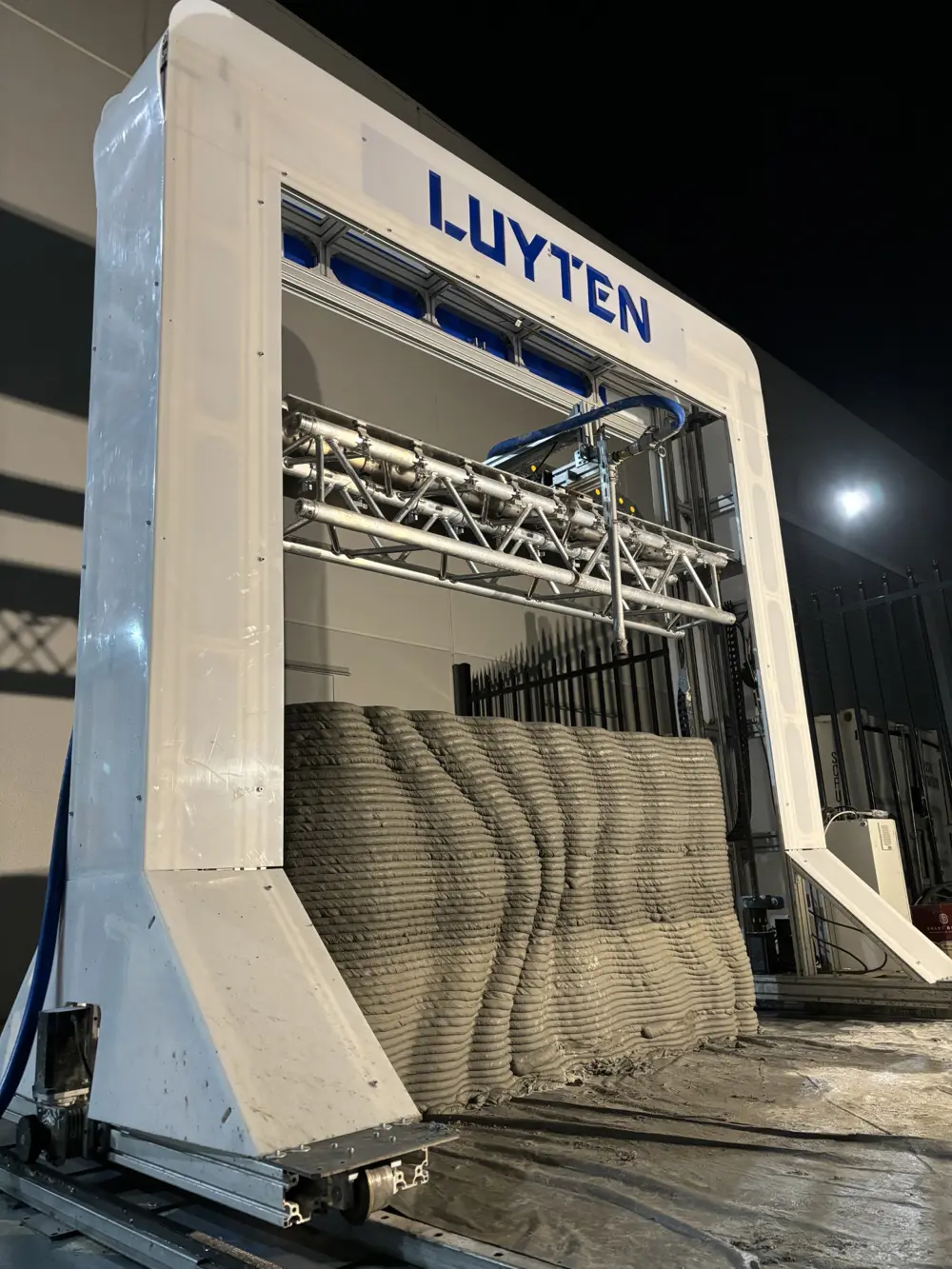 Large gantry‑style 3D printer depositing layered concrete to form a wall section.