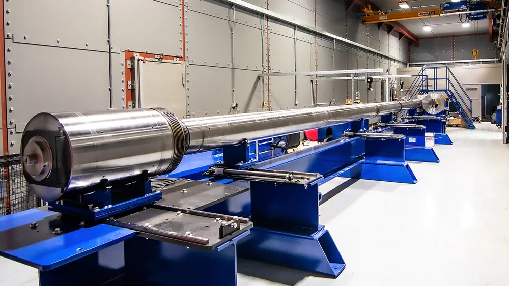 A large silver steel pole, which is wider at one end, sits on a blue steel rack in an industrial setting