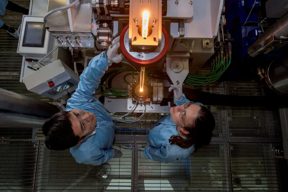 An aerial show of two scientists in a lab controlling the fibre preform being fed into a furnace.