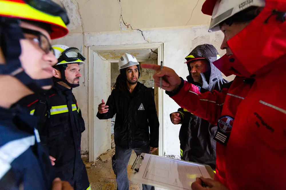 Rescue personnel wearing helmets and protective gear gather inside a damaged building, discussing plans with one person holding a clipboard.