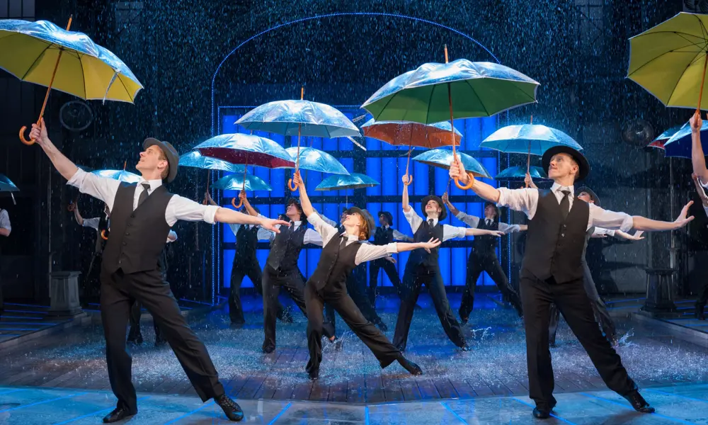Dancers on a stage performing in the "singin' in the rain" musical, pointing umbrellas towards the ceiling. Water is falling down on them and they are splashing in puddles of water as they dance. 