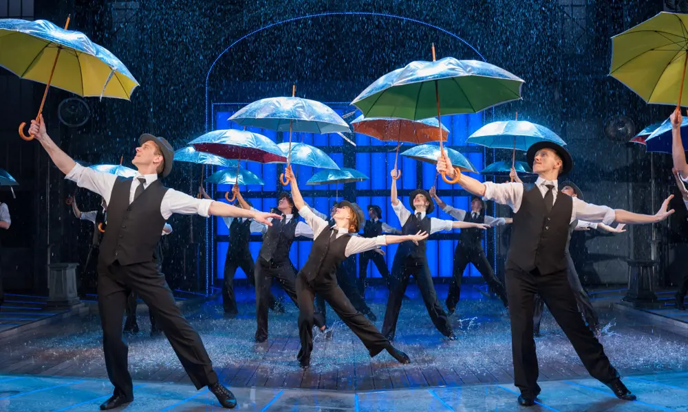 Dancers on a stage performing in the "singin' in the rain" musical, pointing umbrellas towards the ceiling. Water is falling down on them and they are splashing in puddles of water as they dance. 
