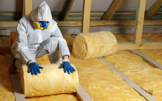 A person wearing a white PPE suit and blue gloves, unrolling insulation in an attic.