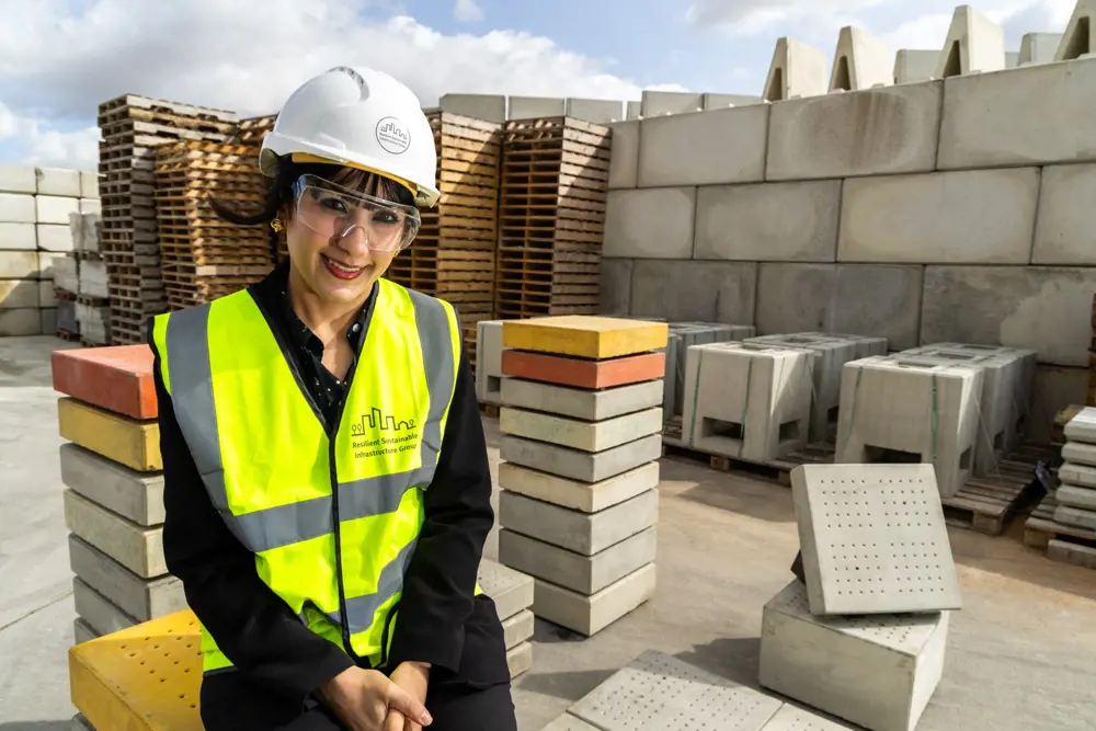 A woman in a high vis jacket and hard hat sits on a pile of slabs of concrete with a grid of small holes