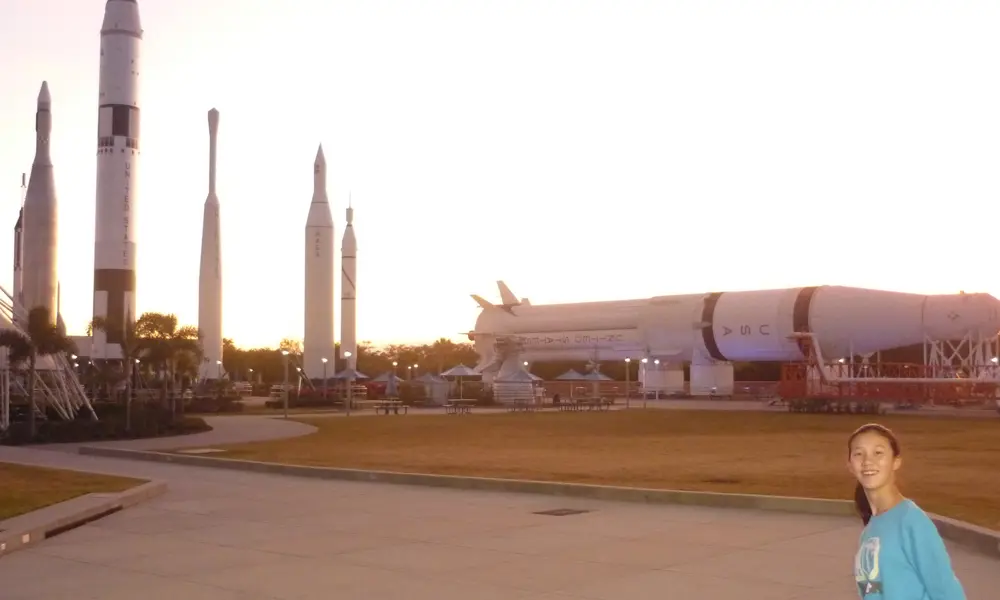 Several large rockets and a horizontal launch vehicle are displayed outdoors at a space centre at sunset, with paved walkways, seating areas and lighting in the foreground; a person stands at the edge of the frame.