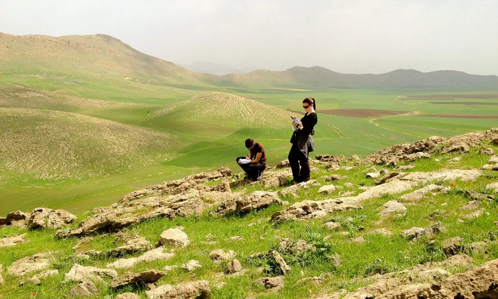 Two people stand on the side of a hill writing on clipboards with other hills in the background