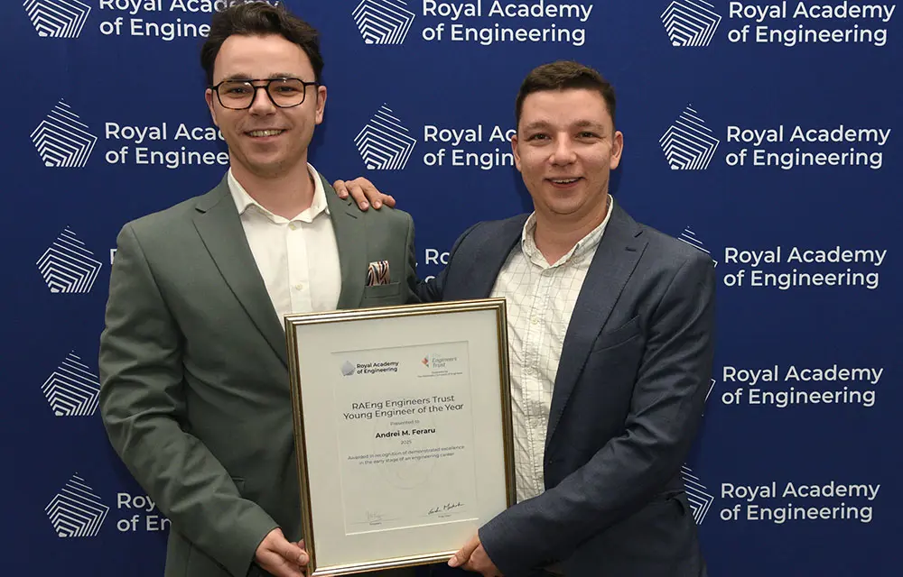 Two people standing in front of a Royal Academy of Engineering backdrop, holding a framed award certificate together.