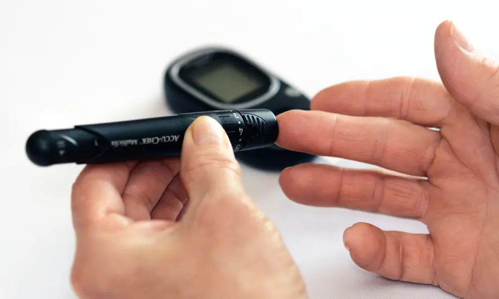A pen-shaped device taking a pinprick of blood from someone's middle finger with a handheld sensor screen in the background.