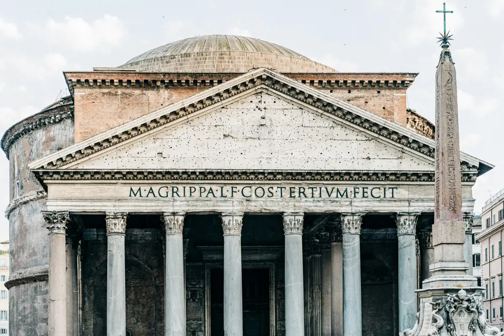 The Pantheon in Rome from outside, with its dome just seen and a bright cloudy sky behind it.