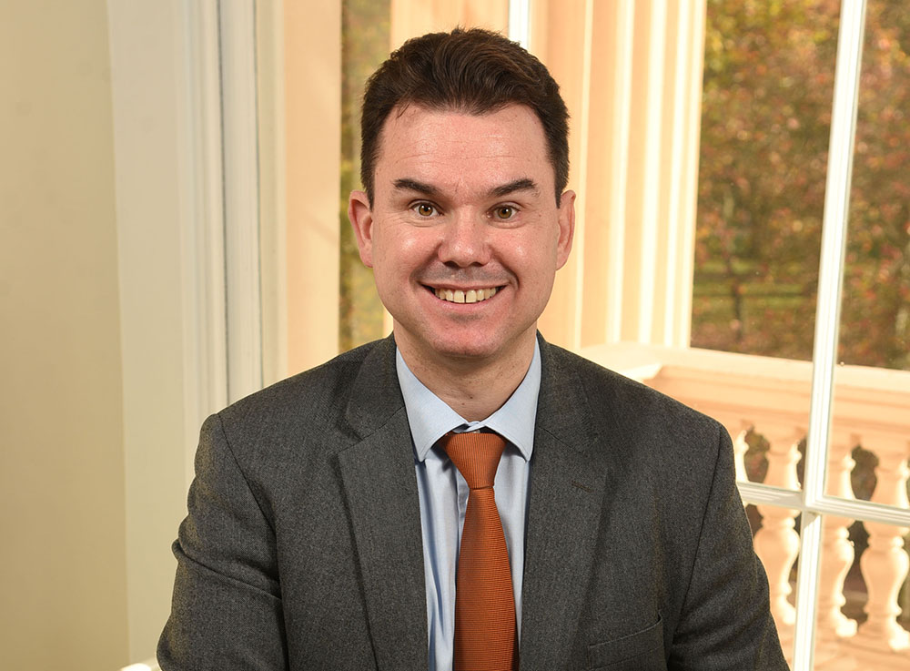 A man with brown short hair wearing a suit jacket and tie smiles at the camera 