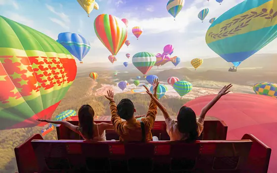Three children sit in a ride carriage with their backs to the camera in front of a simulation of colourful hot air balloons on a large screen 
