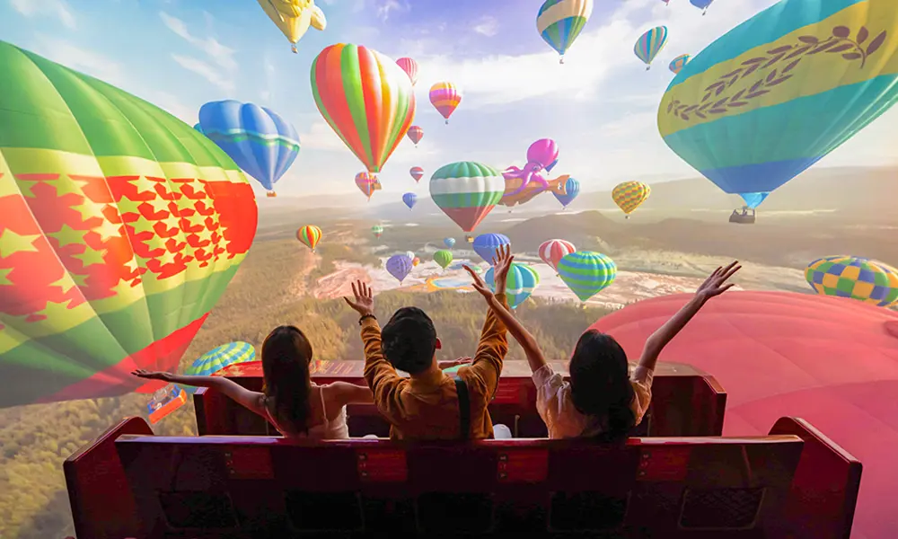Three children sit in a ride carriage with their backs to the camera in front of a simulation of colourful hot air balloons on a large screen 