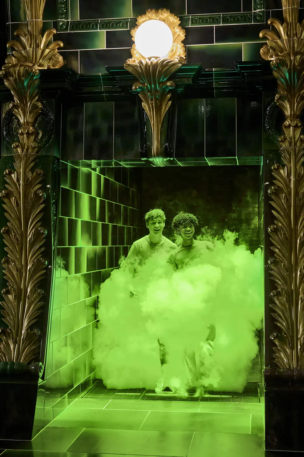 Two teenage boys smile while standing inside a black-tiled fireplace surrounded by green smoke