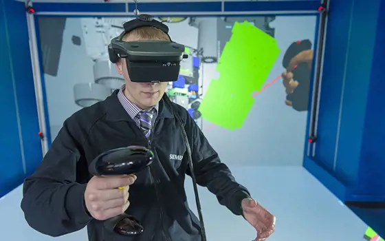 A person with a siemens jacket on wearing a virtual reality headset and a handheld controller to practise engineering.