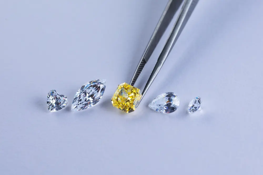 Five synthetic diamonds of different shapes on a white background, with a pair of tweezers picking up a yellow, square diamond in the centre.
