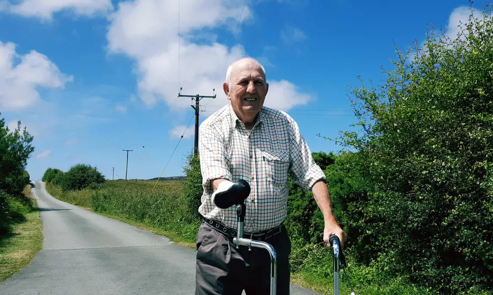Eilian walking on a road lined with green hedges on a sunny day. He is using his adapted three-wheeled rollator.