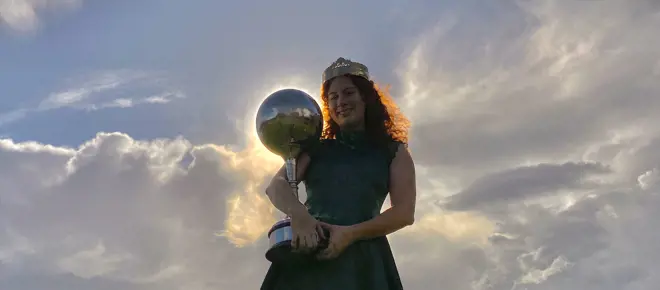 A woman with curly hear, wearing a crown and holding a trophy topped with a large sphere, that is positioned in front of the sun so there is the halo of light around it.