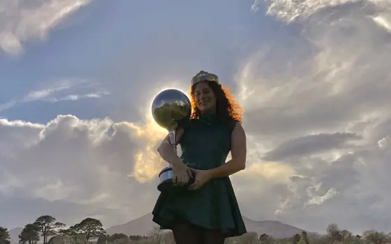A woman with curly hear, wearing a crown and holding a trophy topped with a large sphere, that is positioned in front of the sun so there is the halo of light around it.