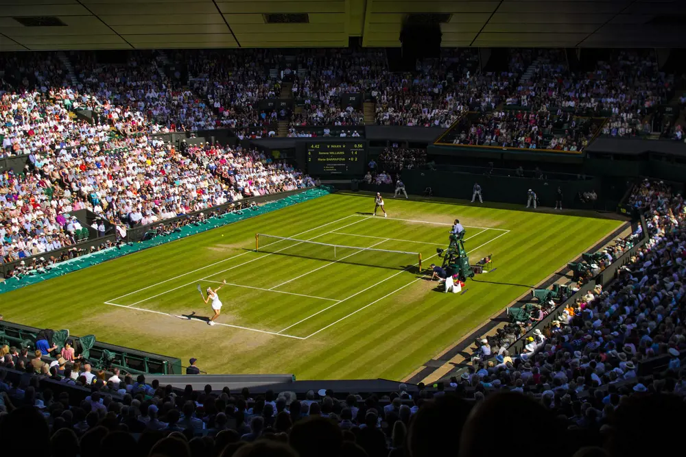 Two women dressed in white play tennis on a green grass court, watched by a full crowd and an umpire