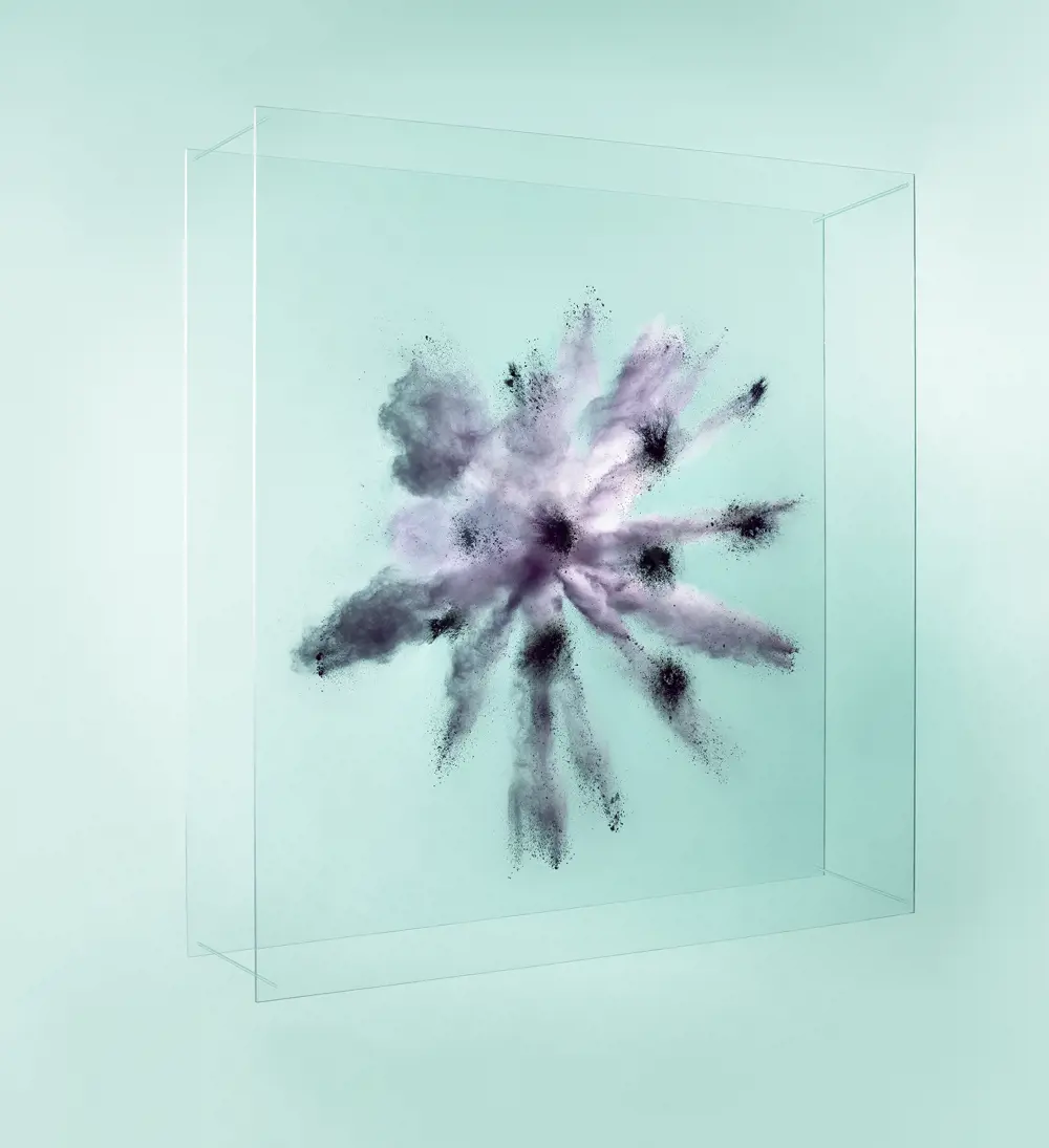 Black-grey powder explodes between two acrylic, transparent sheets, against a light turquoise background