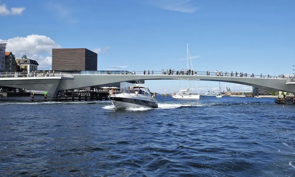 A boat driving under the Inner Harbour Bridge in Copenhagen. People are walking across the bridge. 