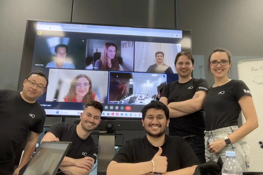 A grinning team of four men and a woman standing in front of a big screen showing a video call with more participants online.