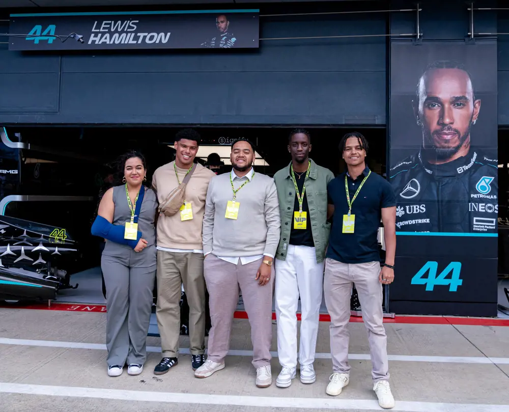 Five people with yellow VIP lanyards posing for a group photo next to a large portrait of Sir Lewis Hamilton at the Mercedes garage