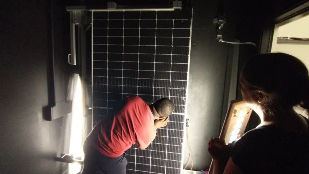 Two people in a dimly lit room examine a solar panel mounted upright, illuminated by bright lamps used to simulate sunlight during testing.
