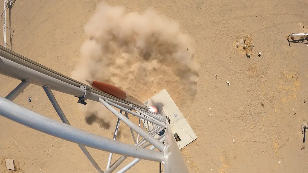 A rocket launching from a metal gantry in a desert, with dust and smoke below.