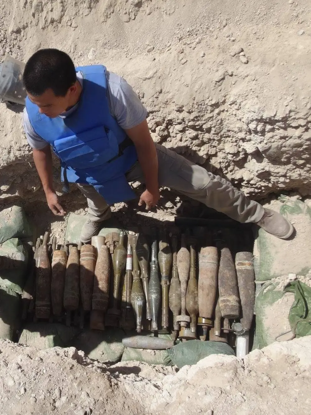 A man wearing a blue vest stands in a hole that has been dug for a demolitions, which the hole filled with explosive ordnance.