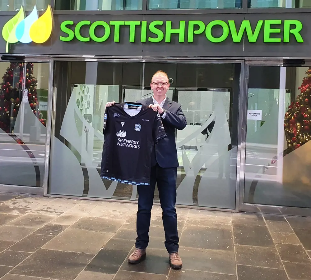 A man standing in front of an office with a "Scottish Power" sign on, holding up a football shirt with "SP Power Networks" in the position of the sponsor logo.
