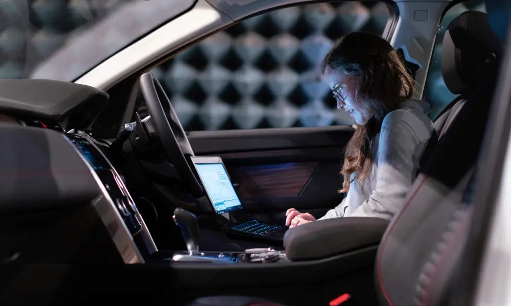 A female electronics engineer runs tests on a laptop while sitting in the driver's seat of a car.