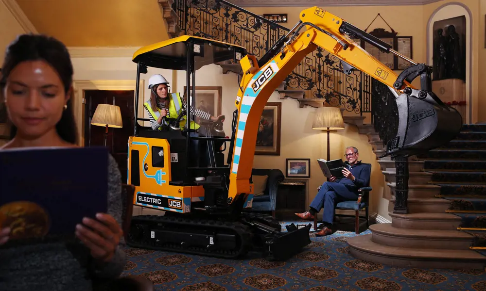 An electric JCB placed inside of a house, with two people reading books around it to demonstrate it's quietness. 