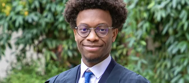 A man with an afro and glasses smiles for a photo, wearing a suit and matriculation gown to enrol at the University of Cambridge.