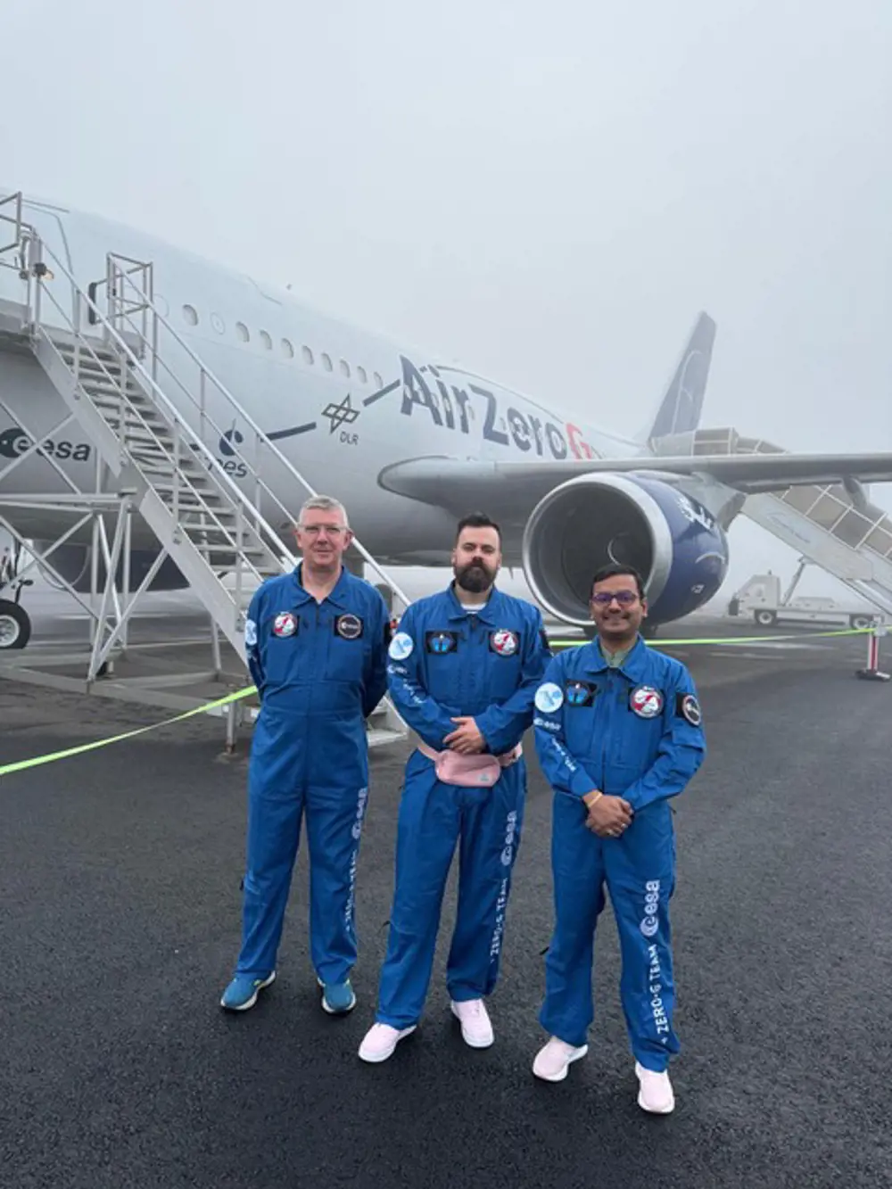 Three men in blue boilersuits posing for a photograph in front of an A310 jet on a misty day.