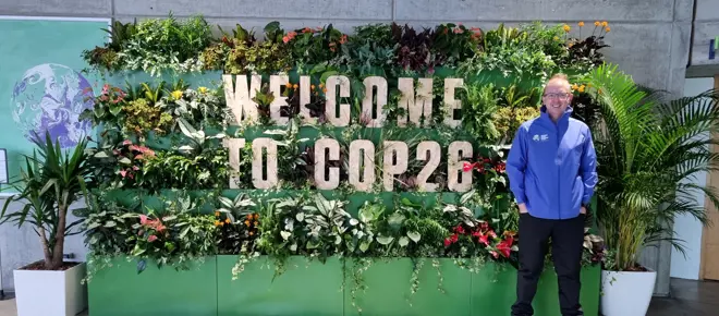 A man standing in front of a display that says "Welcome to COP26" on several shelves full of different plants.
