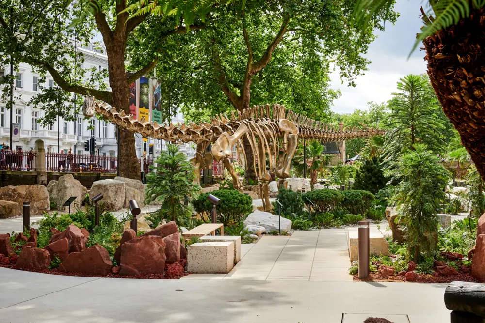 On a sunny day, the sauropod sculpture stands surrounded by plants in a garden.