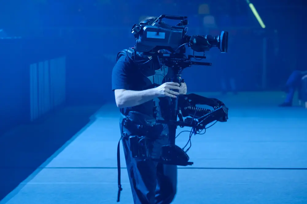 A camera operator using a Steadicam stabiliser rig to hold a large video camera in a blue-lit indoor setting, possibly a stage or event space.
