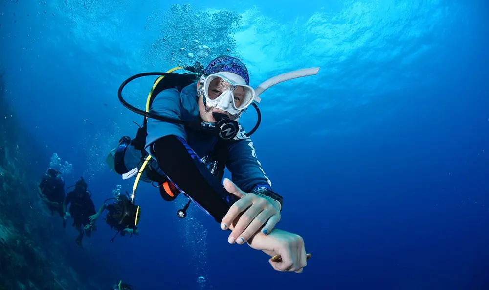 A scuba diver underwater extending an arm forward, with other divers visible in the background against a deep blue ocean.