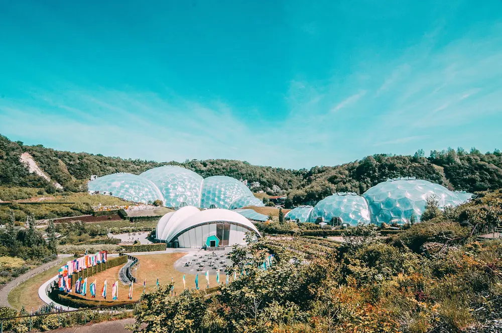 A sweeping outdoor view of the Eden Project, showing several large white geodesic domes set among green hills and landscaped gardens under a bright blue sky.